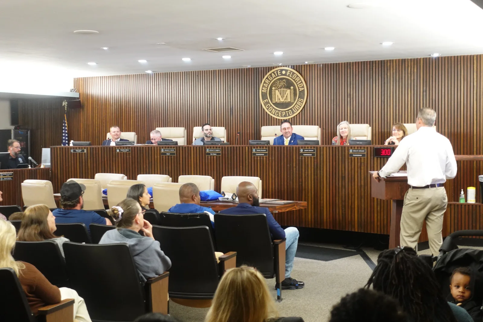 A live City of Margate, Florida council meeting in the wood-paneled chamber, with commissioners at the dais and residents in the audience, in a room outfitted with Full Moon Creative government AV